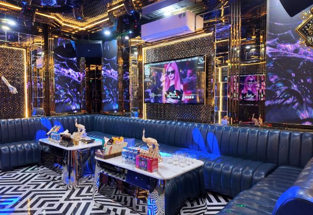 Phòng hát Karaoke Royal 97 Cầu Giấy với sofa da xanh chạy dọc tường, bàn đá, màn hình lớn và tường LED ánh vàng