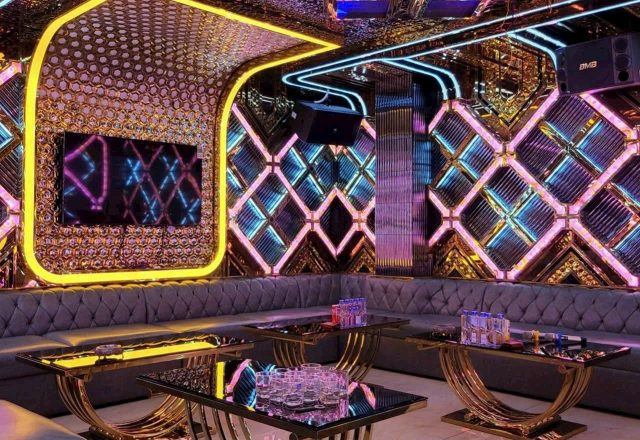 Phòng hát tại Karaoke Tiptop có tường LED hình thoi hồng xanh, sofa xám bọc nút, bàn kính chân vàng và loa treo