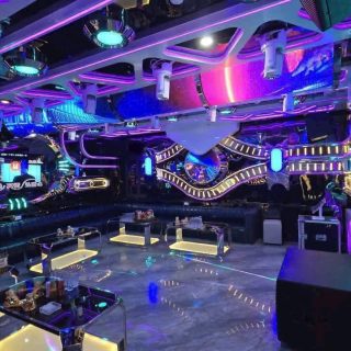 Phòng karaoke rộng của S KTV Club, đèn neon tím xanh, sofa dài quanh tường và màn hình chọn bài