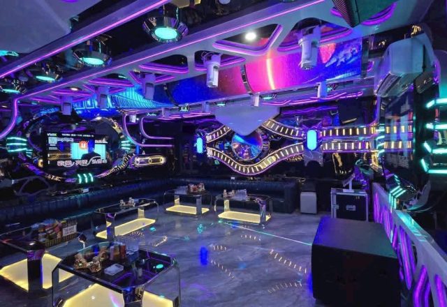 Phòng karaoke rộng của S KTV Club, đèn neon tím xanh, sofa dài quanh tường và màn hình chọn bài