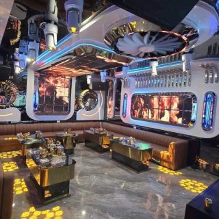 Phòng hát S KTV Club với sofa nâu bọc nệm, bàn mặt kính ánh kim và trần ốp panel trắng phát sáng