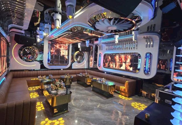 Phòng hát S KTV Club với sofa nâu bọc nệm, bàn mặt kính ánh kim và trần ốp panel trắng phát sáng